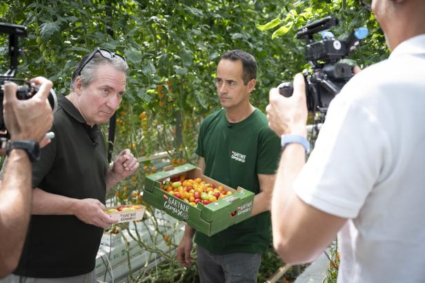 Zwei Männer in einem Gewächshaus, einer hält eine Kiste mit Tomaten, der andere eine Schale mit Tomaten, zwei Kameraleute filmen sie.
