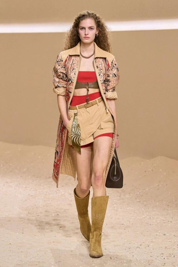 Model läuft auf dem Laufsteg in roter Bandeau-Top, beiger Shorts, gemustertem Mantel und hohen Stiefeln.