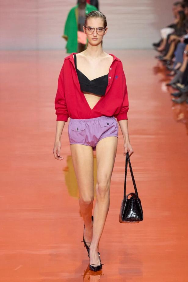 Model auf dem Laufsteg mit roter Bluse, schwarzem Oberteil, lilafarbenen Shorts und schwarzer Handtasche.