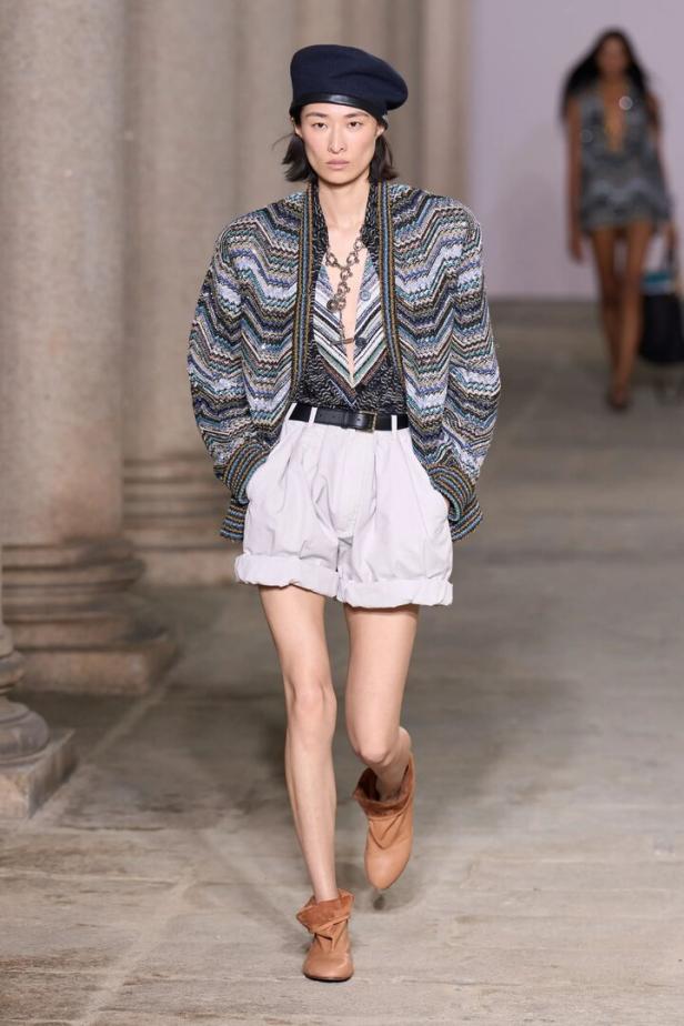 Model auf dem Laufsteg mit gemustertem Blazer, weißen Shorts, Baskenmütze und Stiefeln.