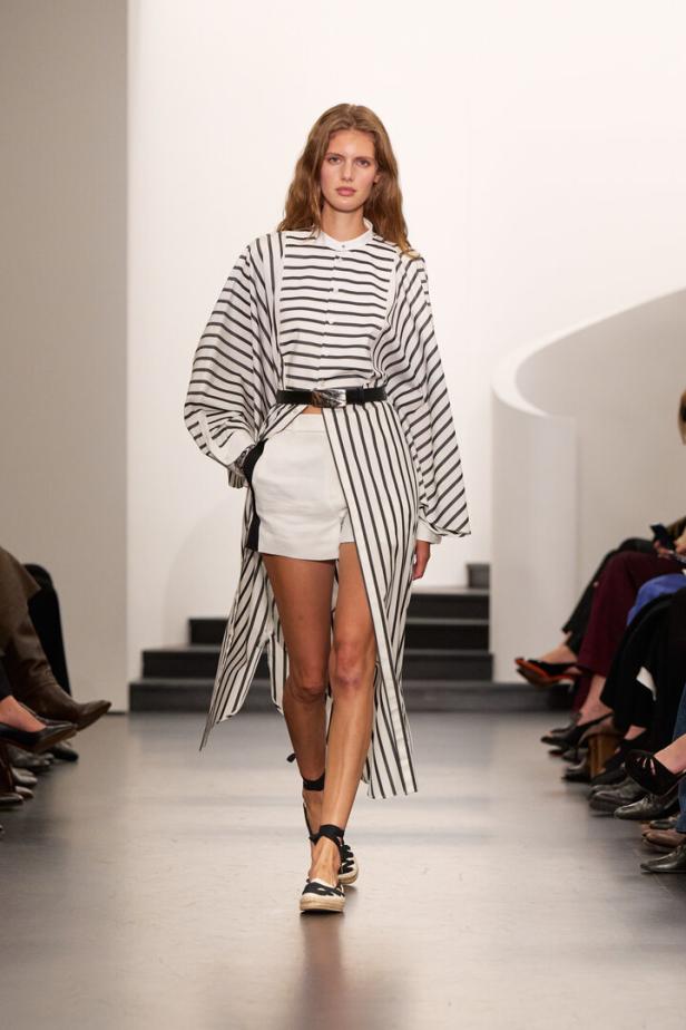 Model auf Laufsteg in gestreiftem Oversize-Hemd, weißen Shorts und Sandalen mit Gürtel.