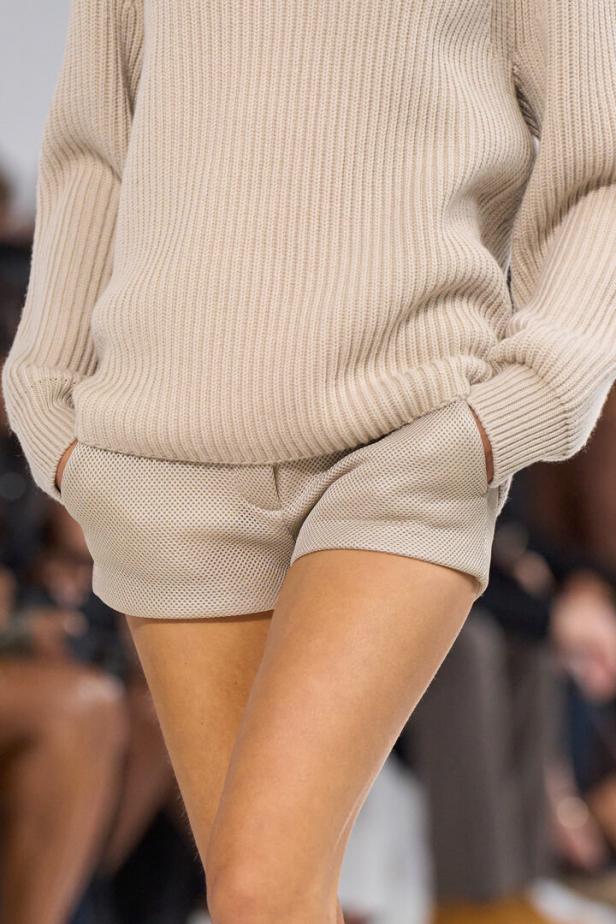 Eine Person trägt einen beigen, gerippten Strickpullover zu kurzen, hellen Shorts mit Netzstruktur.