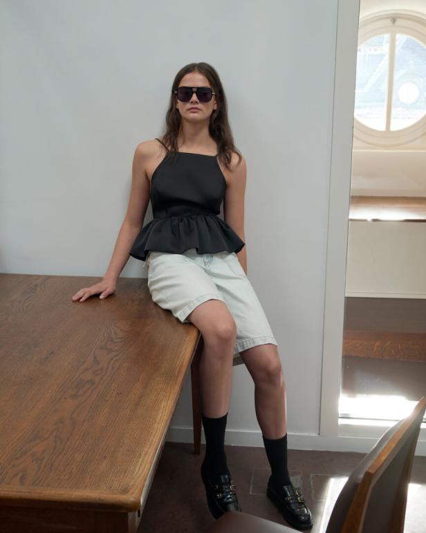 Junge Frau mit Sonnenbrille sitzt in schwarzem Top und hellem Shorts auf einem Holztisch.