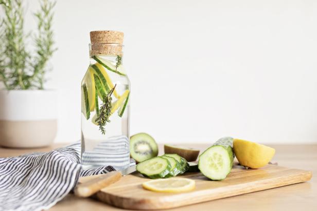 Glasflasche mit Zitronen-, Gurken- und Kräuterscheiben, daneben geschnittenes Obst auf einem Holzbrett.