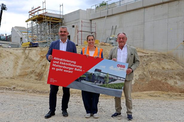 Drei Personen stehen auf einer Baustelle vor einem großen Bahn-Projektplakat.