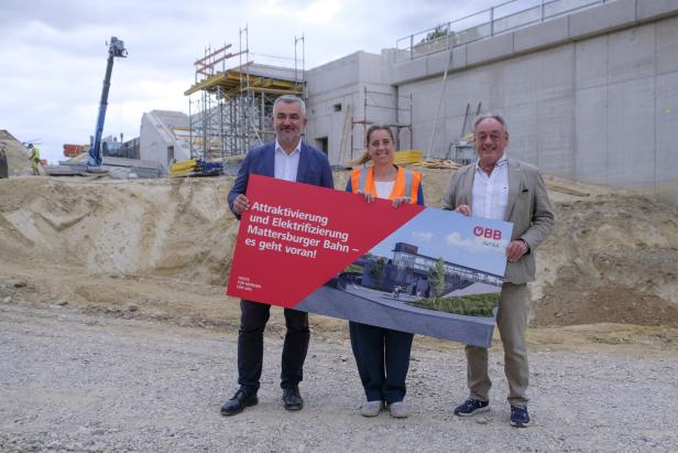 Drei Personen halten ein ÖBB-Infra-Schild auf einer Baustelle für die Attraktivierung und Elektrifizierung der Mattersburger Bahn.