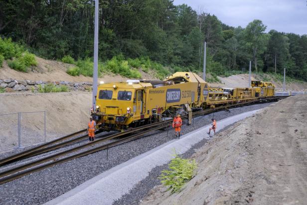 Gelbe Gleisbaumaschine mit Bauarbeitern auf einer Bahnstrecke im Wald.