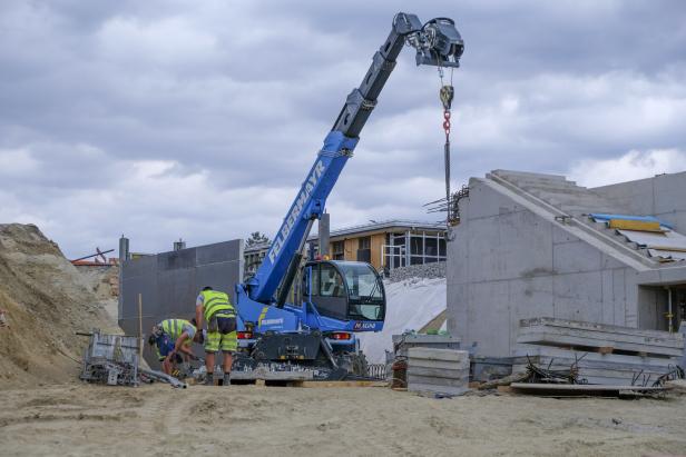 Baustelle mit Kranarm, zwei Arbeitern und Betonbauteilen vor einem Rohbau.