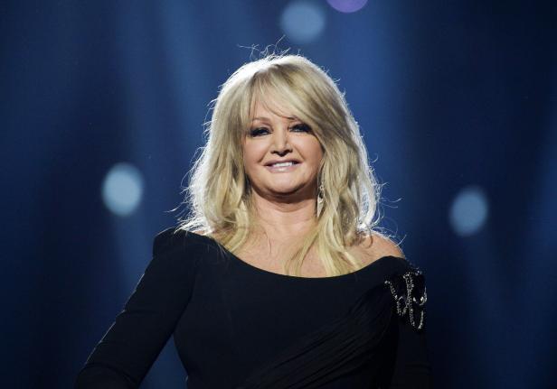 Bonnie Tyler lächelt vor einem dunklen, blau beleuchteten Hintergrund in die Kamera.