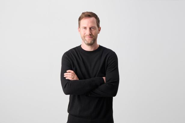 Mann mit schwarzem Pullover steht vor hellem Hintergrund und verschränkt die Arme.