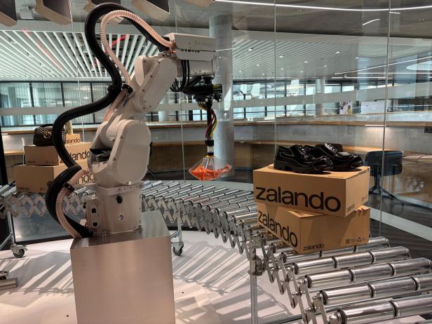 Industrie-Roboter in Lagerhalle packt Schuhe auf ein Förderband neben Zalando-Kartons.