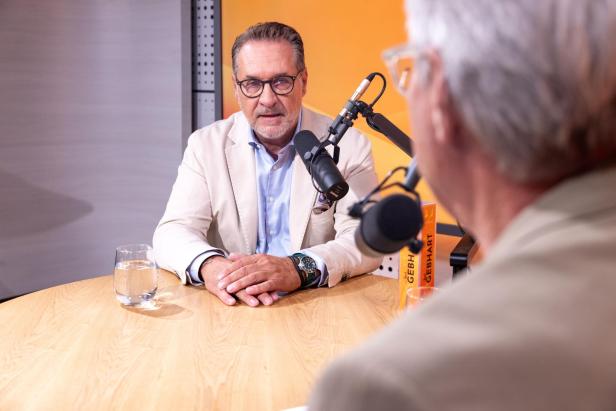 Ein Mann mit Brille und hellem Sakko sitzt an einem Holztisch vor einem Mikrofon und führt ein Gespräch in einem Studio.