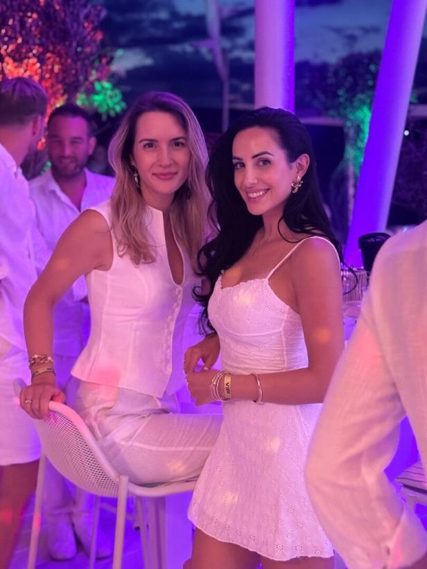 Zwei Frauen in weißen Outfits lächeln bei einer Party im violetten Licht.