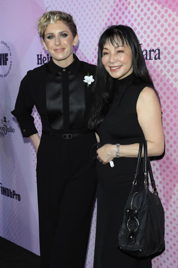 Noriko Watanabe und Ari Wegner in schwarzer Kleidung stehen lächelnd vor einer rosa gemusterten Wand mit Logos wie IMDbPro.