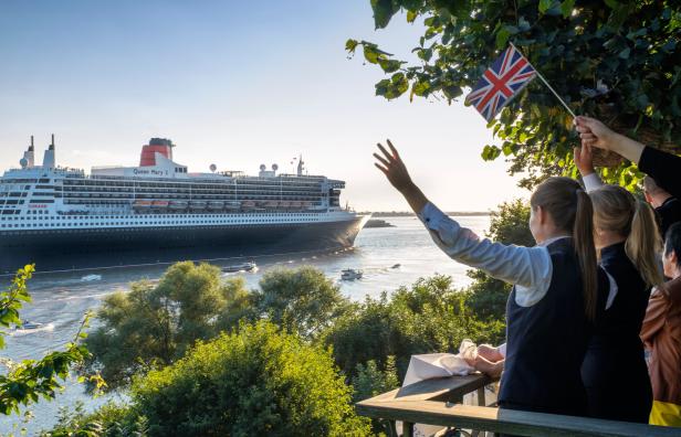 Menschen winken einem vorbeifahrenden Kreuzfahrtschiff „Queen Mary 2“ zu, eine Person hält eine britische Flagge.