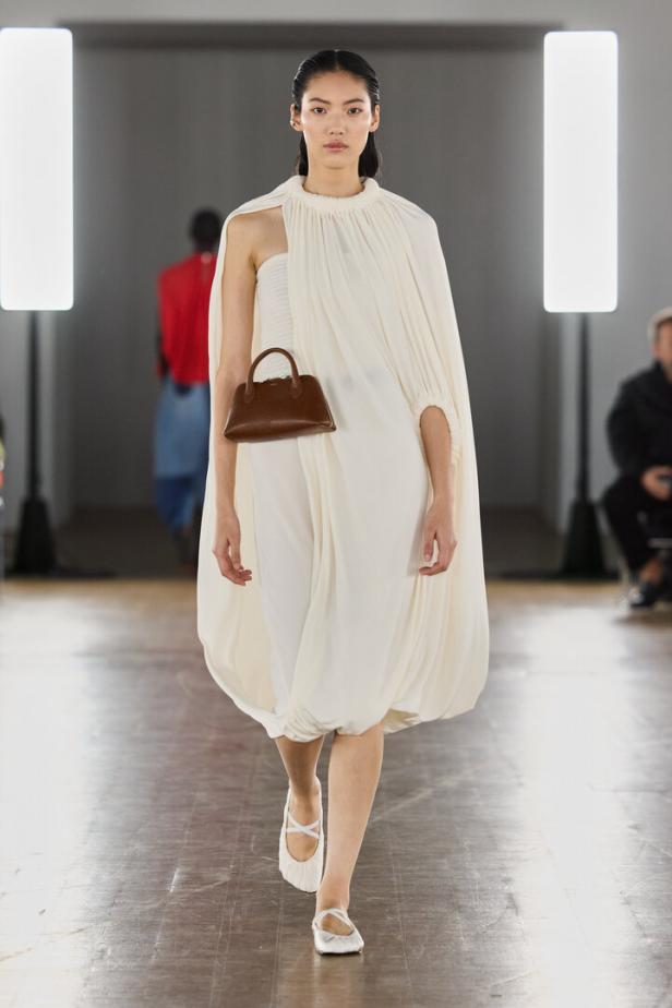 Model auf dem Laufsteg in fließendem weißen Kleid mit brauner Handtasche und Ballerinas.