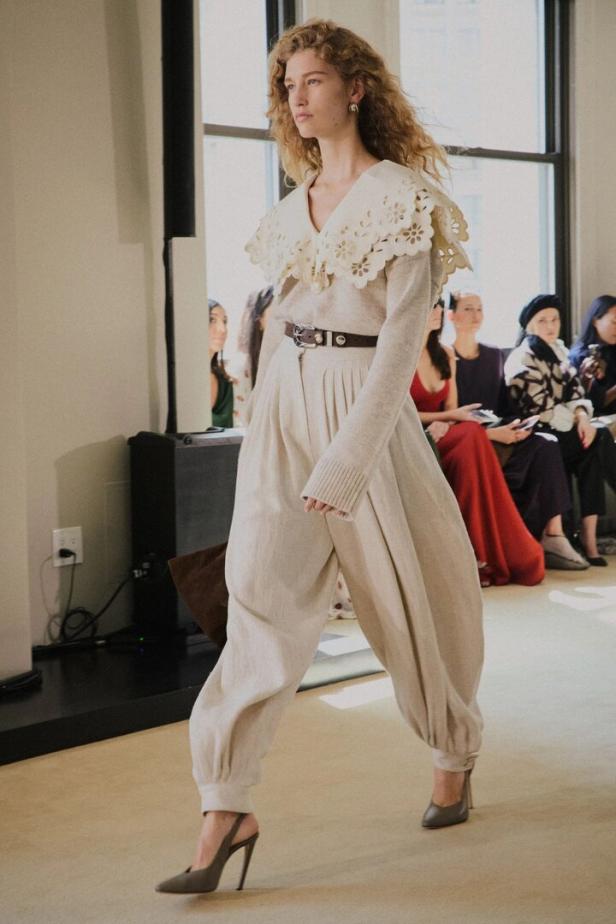 Model auf dem Laufsteg in cremefarbenem Outfit mit Spitzkragen und hoher Taille, vor sitzendem Publikum.
