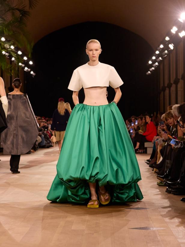 Model auf dem Laufsteg mit weißem Crop-Top und voluminösem grünen Rock, flankiert von Publikum.