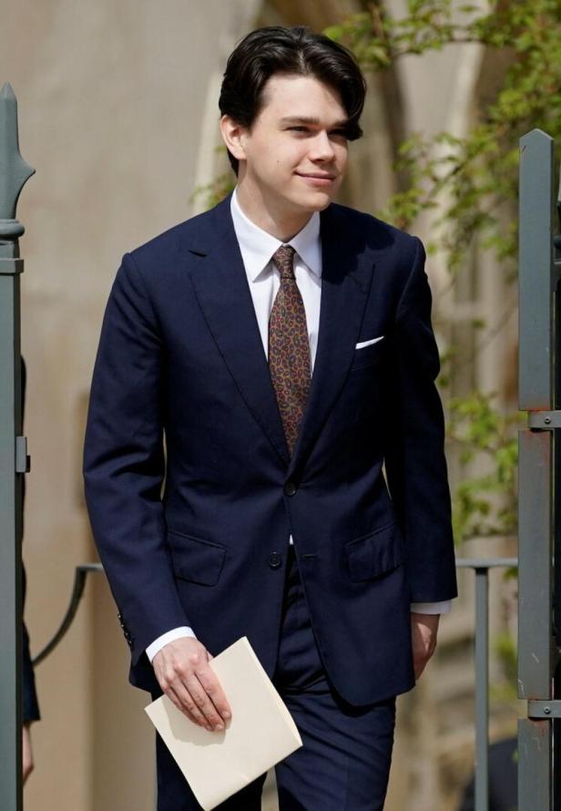 Samuel Chatto mit dunklem Haar trägt einen dunkelblauen Anzug, ein weißes Hemd und eine gemusterte Krawatte und hält ein helles Heft in der Hand.