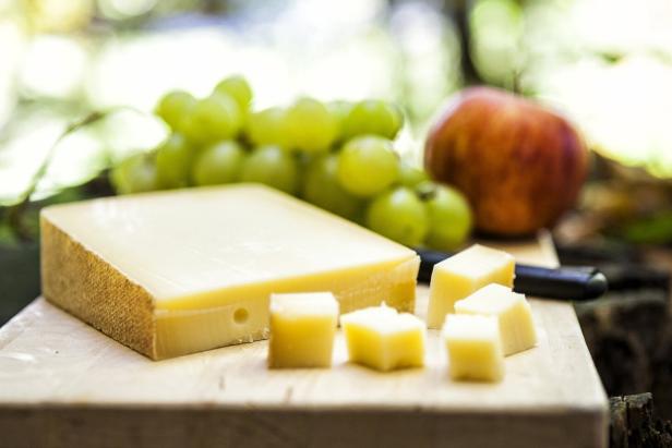 Ein Stück Käse mit gewürfelten Stücken liegt neben grünen Weintrauben und einem Apfel auf einem Holzbrett.