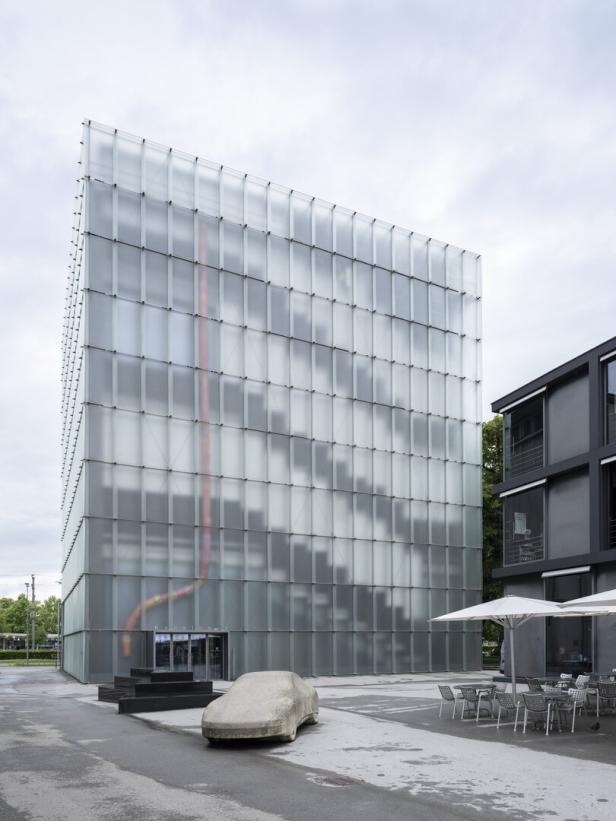 Modernes Glasgebäude mit großem Eingangsbereich, davor eine skulpturale Autoinstallation und Sitzgruppe im Innenhof.