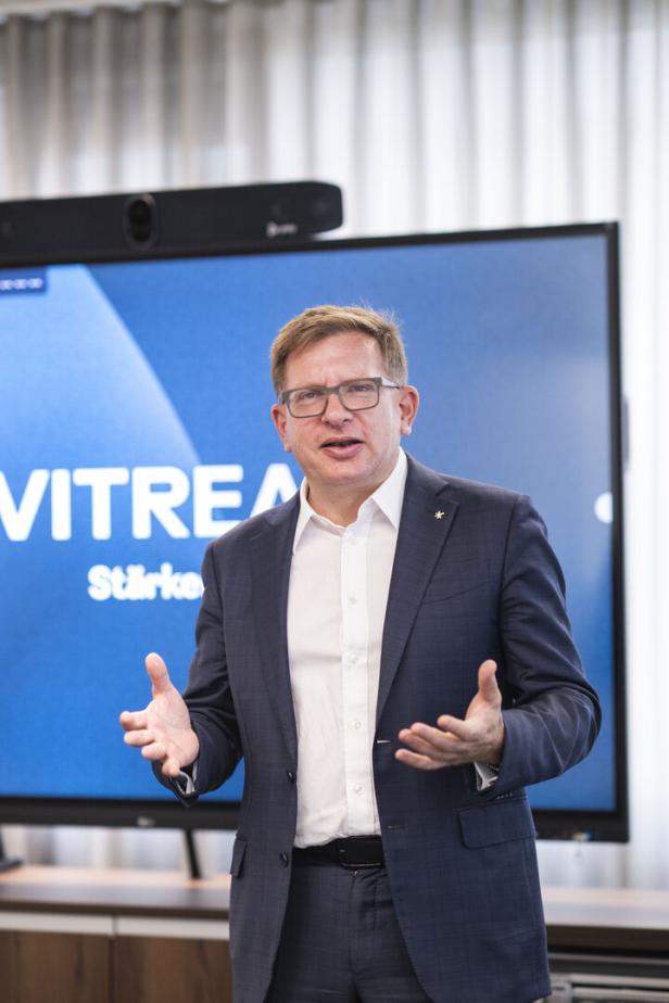 Mann im Anzug spricht vor einem großen Bildschirm mit blauem Hintergrund.