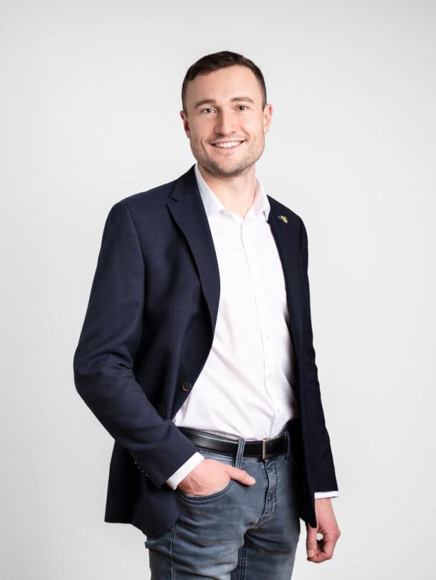 Lächelnder Mann im dunkelblauen Sakko, weißem Hemd und Jeans vor hellem Hintergrund.