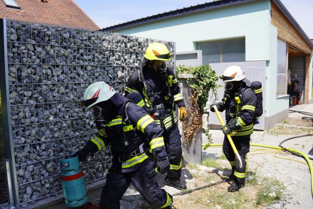 Drei Feuerwehrleute in Schutzanzügen arbeiten neben einem Haus mit Schläuchen und Gasflasche.