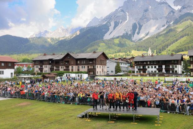 Menschenmenge bei einer Bergveranstaltung vor alpiner Kulisse mit schneebedeckten Gipfeln und Bühne.