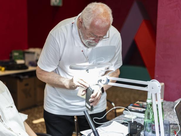 Ein älterer Mann repariert unter einer Lupe ein Gerät am Tisch.