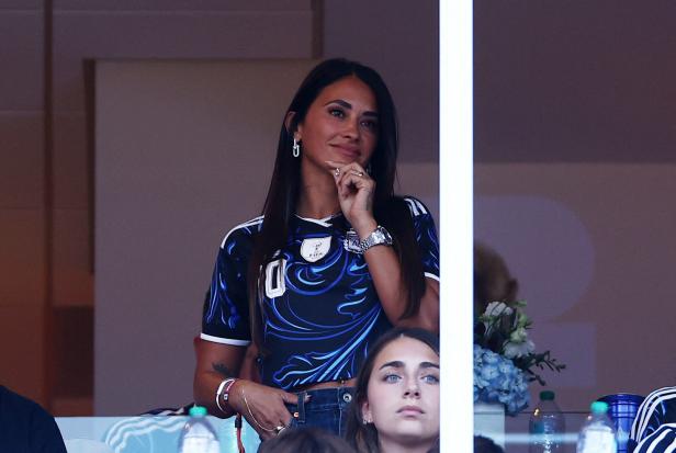 Antonela Roccuzzo trägt ein blau-schwarzes Trikot und schaut lächelnd von einer Tribüne aus zu.