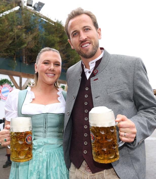 Harry Kane und seine Frau Katie Goodland in traditioneller bayerischer Tracht halten jeweils einen Paulaner-Maßkrug in die Kamera.