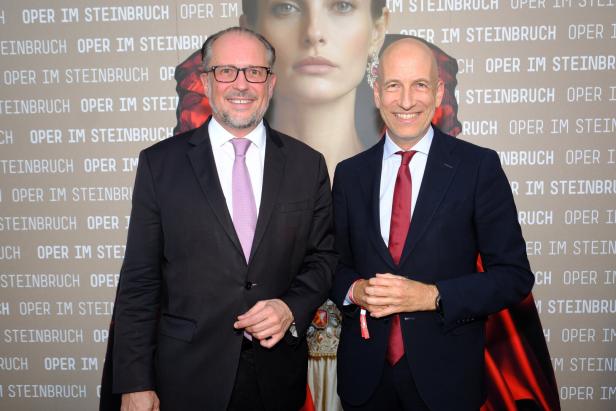 Zwei Männer im Anzug posieren vor einer Opern-Premierenwand mit der Aufschrift „Oper im Steinbruch“.