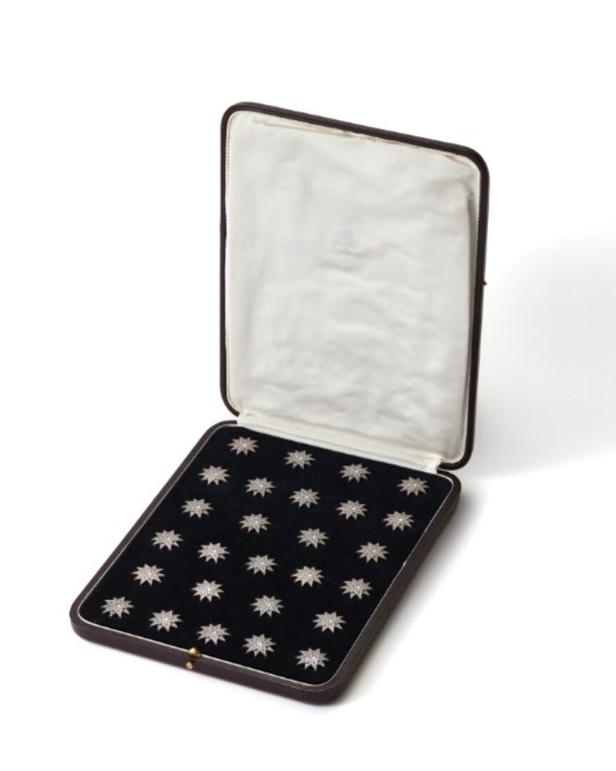 Ein offenes, dunkelbraunes Etui mit weißem Innenfutter präsentiert auf schwarzem Samt zahlreiche kleine, sternförmige Schmuckstücke.