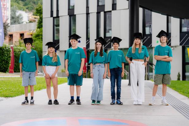 Sechs Kinder mit Doktorhüten und türkisfarbenen Shirts stehen draußen vor einem modernen Gebäude.