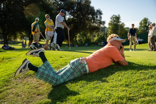 Mann im Golfoutfit liegt auf dem Grün und telefoniert, während andere Spieler in der Nähe stehen.