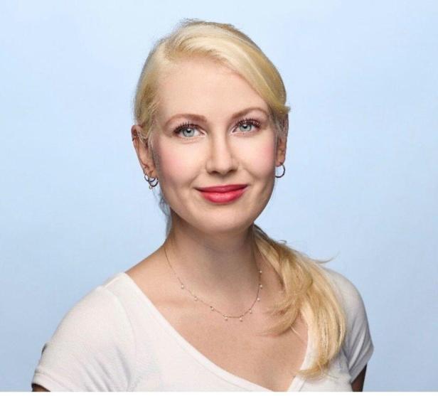 Porträt einer blonden Frau mit Zopf, hellem Oberteil und dezentem Schmuck vor hellblauem Hintergrund.