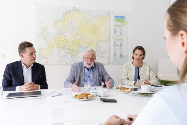 Drei Personen diskutieren an einem Tisch in einem hellen Besprechungsraum vor einer Europakarte.