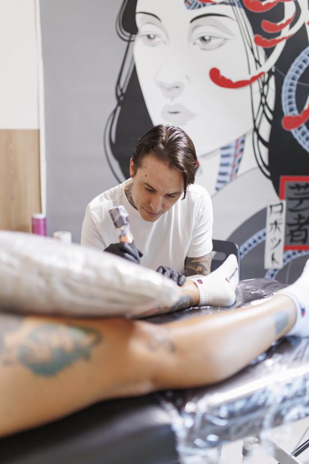 Ein Tätowierer arbeitet konzentriert an einem Kunden, während im Hintergrund eine große Frauenillustration hängt.