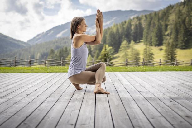 Frau macht Yoga auf einer Holzterrasse mit Berg- und Waldkulisse.