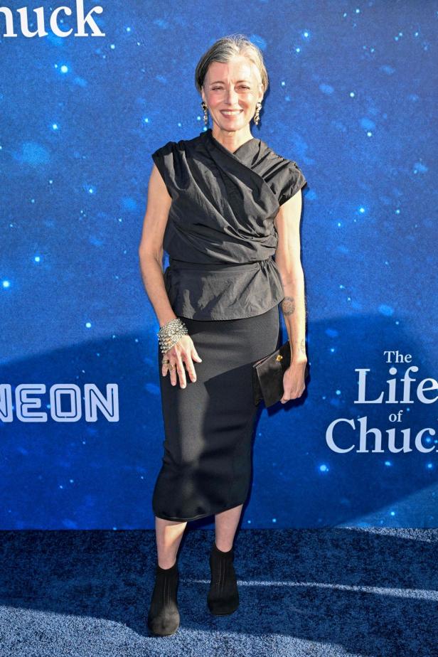 Mia Sara mit kurzem, grauem Haar trägt ein schwarzes Oberteil und einen Rock vor einem blauen Hintergrund mit dem Schriftzug „The Life of Chuck“.