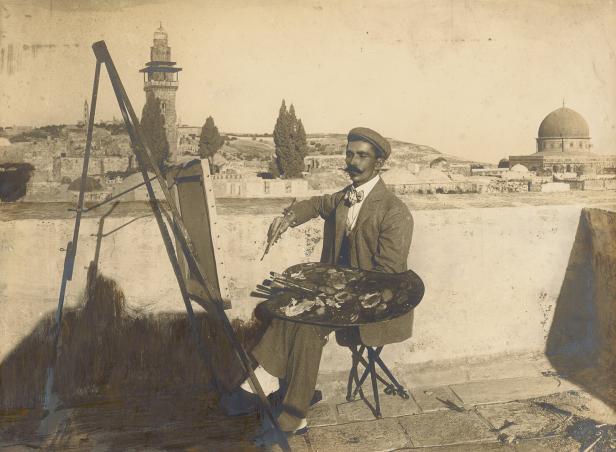 Ein Maler sitzt an der Staffelei mit Paletten und Pinseln vor der Silhouette Jerusalems.