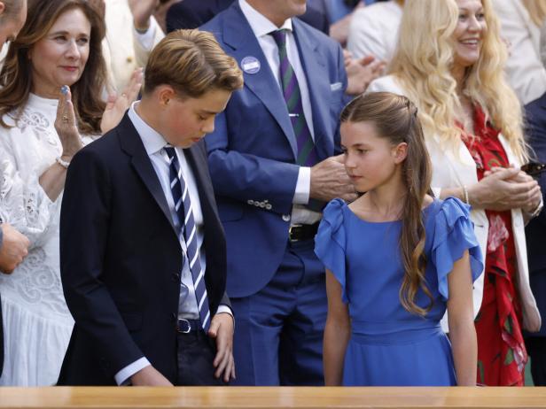 Prinz George und Prinzessin Charlotte stehen nebeneinander und schauen nach unten, umgeben von anderen Personen in festlicher Kleidung.