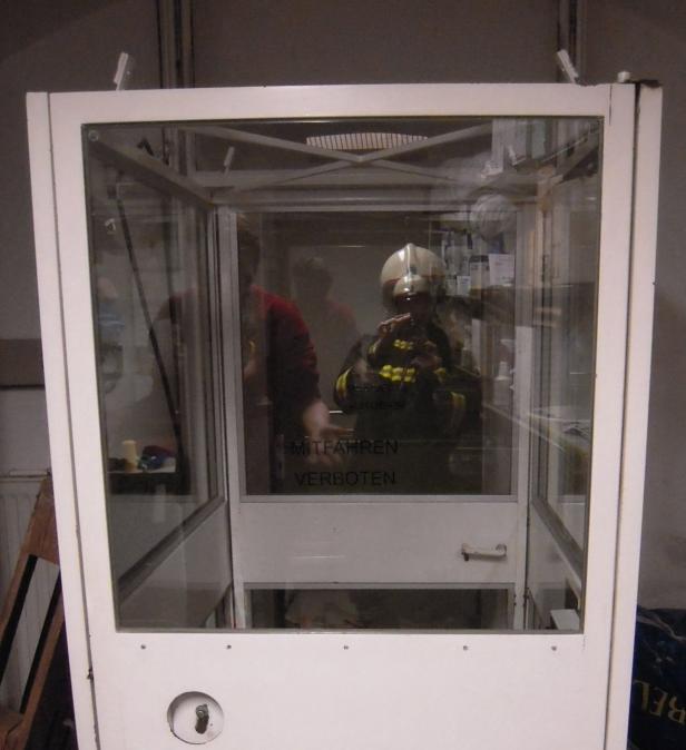 Ein Feuerwehrmann steht in einer gläsernen Kabine, auf der „Mitfahren verboten“ steht.
