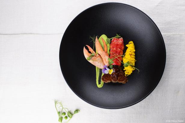 Edles Gourmetgericht mit Hummer, Pasta und Gemüse auf schwarzem Teller, mit essbaren Blüten garniert.