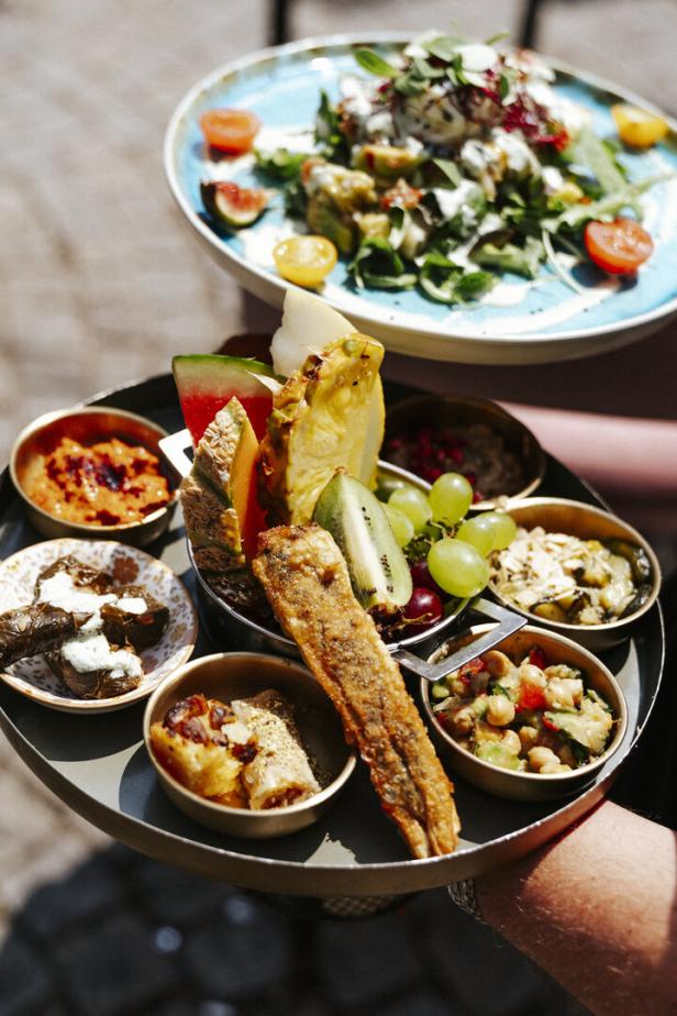 Ein Serviertablett mit verschiedenen Snacks, Obst und Dips wird gehalten, dahinter steht ein Teller mit Salat.