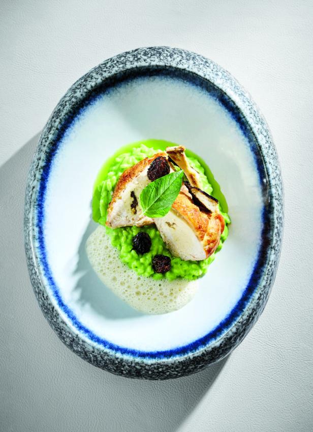 Angebratenes Geflügel auf grünem Risotto mit Kräutergarnitur und Sauce in einer Keramikschale.