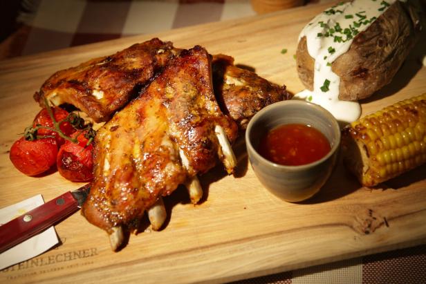 Gegrillte Rippchen mit Tomaten, Ofenkartoffel, Maiskolben und Dip auf einem Holzbrett.