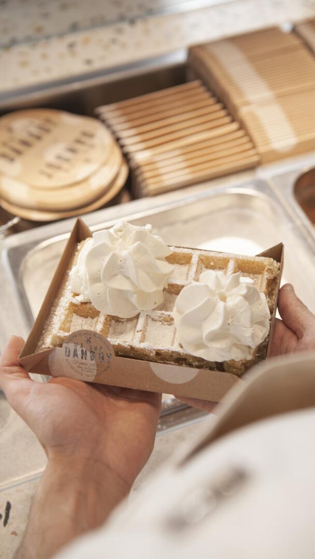 Hände halten eine Waffel in Schachtel mit zwei Sahnehäubchen an einer Desserttheke.