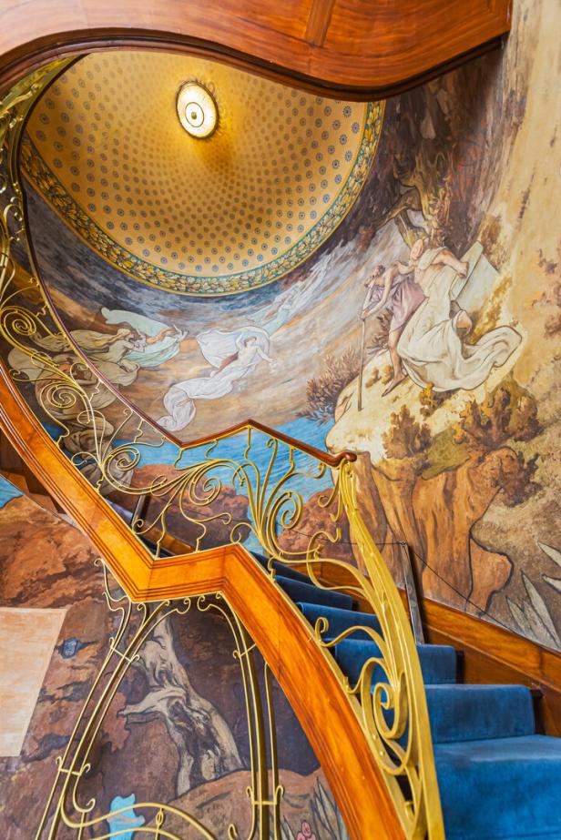Prunkvolle Wendeltreppe mit blauer Stufen und goldenen Geländern vor einer kunstvollen Wand- und Deckenmalerei.